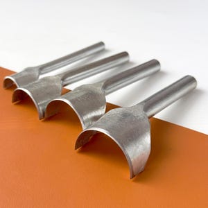 Puede incluir: Cuatro herramientas metálicas para trabajar el cuero con cuchillas curvas, dispuestas en diagonal sobre un fondo blanco y naranja. Las herramientas tienen un filo redondeado y semicircular y un mango largo y cilíndrico. Las herramientas son de metal brillante color plata.