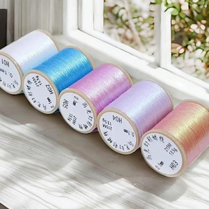 May include: A collection of six spools of iridescent thread in various colors, including white, blue, pink, and peach. Each spool has a label with text and measurements. The spools are arranged in a row on a light-colored surface.