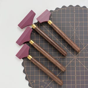 Può includere: Quattro strumenti per la lavorazione della pelle con lame bordeaux, manici in legno scuro e puntali in ottone. Gli strumenti sono disposti diagonalmente su un tappetino da taglio nero con linee a griglia. I manici sono marrone scuro.