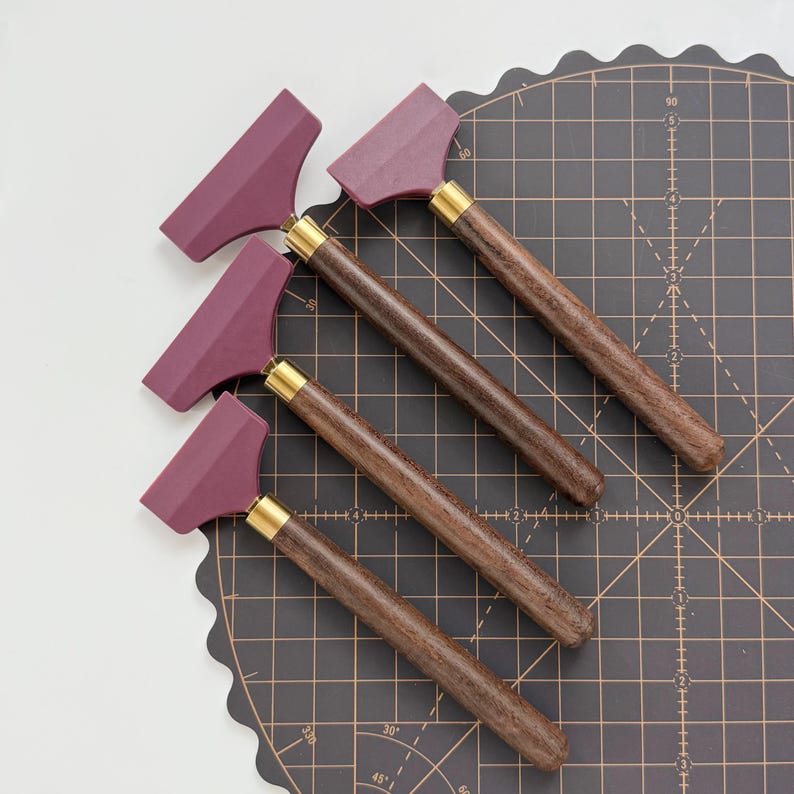 K&ouml;nnte beinhalten: Vier Lederhandwerkzeuge mit weinroten Klingen, dunklen Holzgriffen und Messingzwingen. Die Werkzeuge sind diagonal auf einer schwarzen Schneidematte mit Rasterlinien angeordnet. Die Griffe sind ein sattes, dunkles Braun.