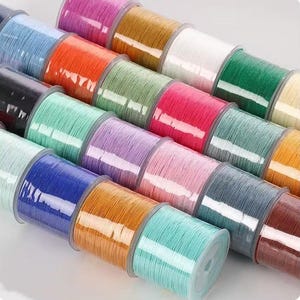 May include: A collection of cylindrical spools arranged in rows, each wrapped with thin, colored cord. The assortment features a vibrant spectrum including blue, orange, green, pink, yellow, white, black, and brown shades in a smooth finish.
