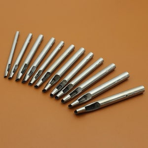 Puede incluir: Una colección de herramientas de perforación de cuero de metal plateado dispuestas en diagonal sobre una superficie marrón. Cada herramienta tiene un cuerpo cilíndrico con un filo biselado y un centro hueco. Las herramientas están numeradas.