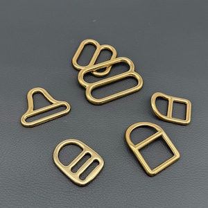 Pode incluir: Uma coleção de peças de ferragens metálicas douradas em várias formas, incluindo argolas em D, laços retangulares e um design triangular. As ferragens são exibidas em uma superfície cinza escuro.
