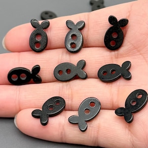 Puede incluir: Una colección de pequeños botones negros con forma de pez. Cada botón tiene dos agujeros para coser y una cola estilizada. Los botones, hechos de un material liso, están dispersos sobre una superficie clara.