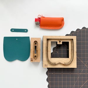 May include: An assortment of leathercraft tools and materials. Includes a teal leather piece, a wooden stamp, a wooden die, and an orange leather lighter case. The wooden die is labeled with "B2".