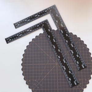 Puede incluir: Escuadras metálicas negras con marcas blancas, que muestran medidas en centímetros. Las reglas están colocadas sobre una esterilla de corte marrón oscuro con un patrón de cuadrícula, ideal para manualidades y medición.
