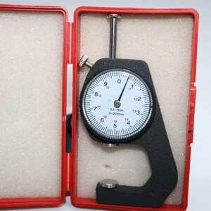 May include: A black thickness gauge with a white circular dial face, featuring black and red numerical markings, a silver needle, and a 0-20mm measurement range, housed inside a red plastic storage case with white foam padding.