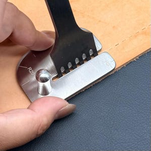 Op de afbeelding: Een roestvrijstalen hoekafrondingsmal voor leer met R5- en R20-markeringen, voorzien van een verticale gleuf voor een zwarte metalen stansbeitel. Het gereedschap is geplaatst op een stuk beige leer voor een nauwkeurige uitlijning.
