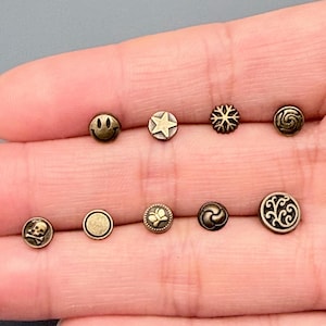May include: A selection of small, antique brass-toned buttons featuring diverse designs such as a smiley face, star, snowflake, swirl, skull, circle, butterfly, and ornate patterns. These decorative buttons are suitable for sewing and crafts.