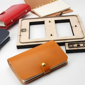 May include: A tan leather notebook with a gold snap closure, a red leather notebook, a blue notebook, an open notebook with gold rings, and a rectangular wooden die-cutting tool with two rectangular cutouts and metal blades.