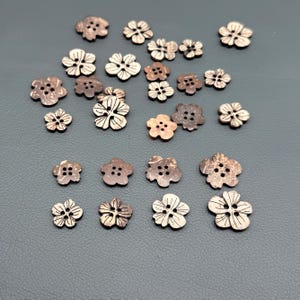 May include: A collection of brown, flower-shaped buttons made from natural material. The buttons vary in size and feature four holes in the center, with some displaying engraved petal lines and others showing a smooth, textured surface finish.