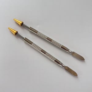 Può includere: Due strumenti argentati per nail art con manici strutturati. Un'estremità presenta un cono dorato, l'altra una piccola spatola piatta. Questi strumenti sono progettati per applicazioni di nail art dettagliate.