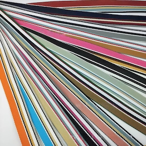 May include: A collection of woven fabric ribbons arranged in a fan pattern. Each ribbon features a distinct solid color including orange, blue, pink, black, green, and tan, accented by thin white and black diagonal stripes along the edges.