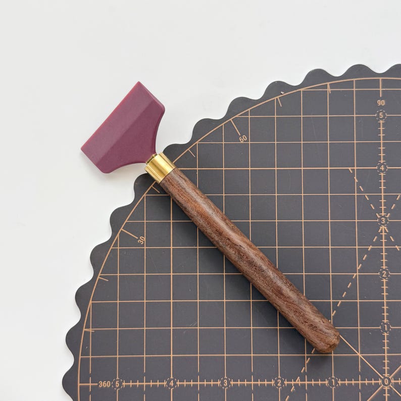 K&ouml;nnte beinhalten: Ein Lederhandwerks-Polierwerkzeug mit einem dunkelvioletten Kopf, einer Messingzwinge und einem dunklen Holzgriff. Das Werkzeug liegt auf einer schwarzen Schneidematte mit Rasterlinien und einem gewellten Rand.