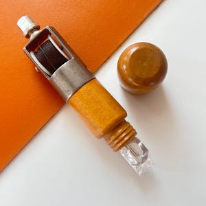 May include: A wooden leather stitching tool with a silver metal frame holding black thread. A small, round wooden knob sits next to the tool. The tool has a clear plastic piece at the end. The background is white and orange.