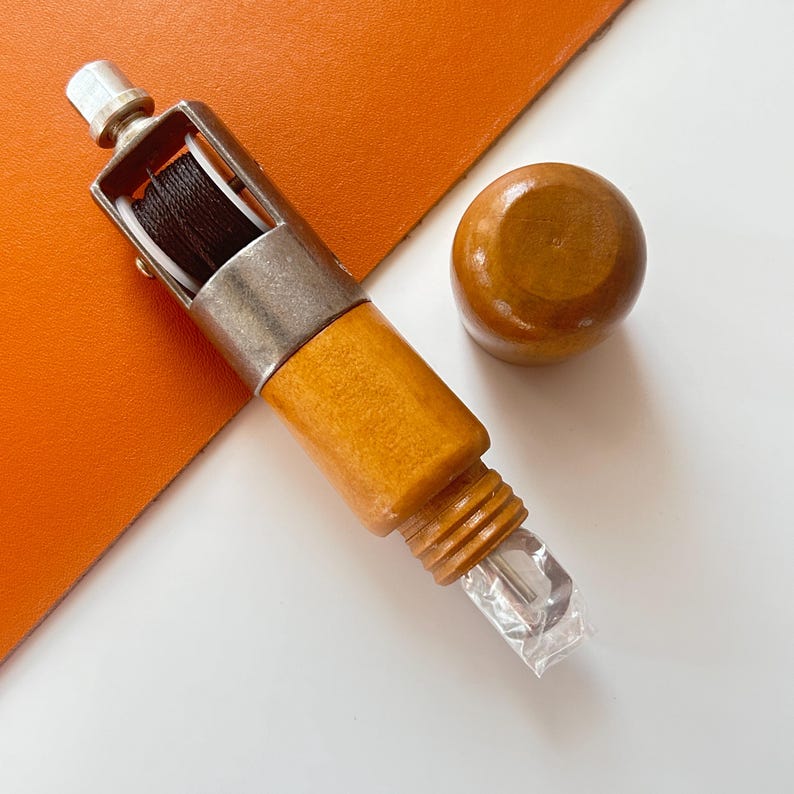 May include: A wooden leather stitching tool with a silver metal frame holding black thread. A small, round wooden knob sits next to the tool. The tool has a clear plastic piece at the end. The background is white and orange.