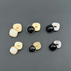 May include: Six square buttons arranged in two rows. The top row features one cream button with a gold back, one black button with a gold back, and one black button with a silver back. The bottom row mirrors this with identical button styles.