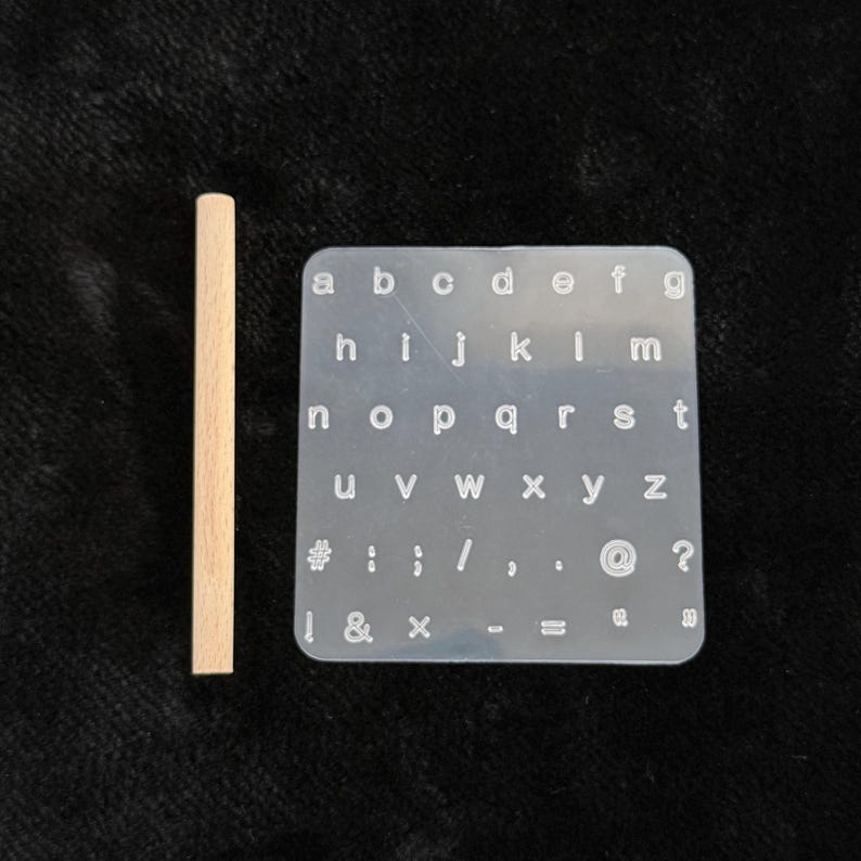 May include: A clear silicone alphabet stamp with lowercase letters and symbols, including numbers, exclamation points, and question marks. A wooden dowel is positioned to the left of the stamp. The stamp is on a dark background.