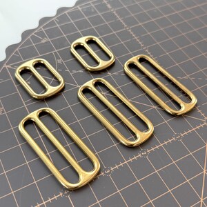 May include: Six gold-coloured rectangular slider buckles with two slots. The buckles are made of a shiny metal and are arranged on a dark grey surface with grid lines. These buckles are likely used for straps or belts.
