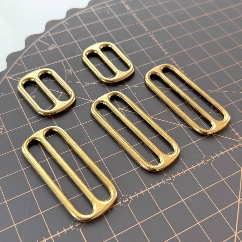 May include: Six gold-coloured rectangular slider buckles with two slots. The buckles are made of a shiny metal and are arranged on a dark grey surface with grid lines. These buckles are likely used for straps or belts.