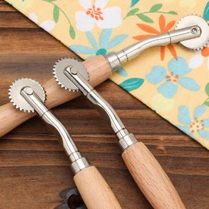 Può includere: Due taglia pasta a rotella con manici in legno e lame in acciaio inossidabile. Le lame hanno un bordo seghettato. Gli strumenti sono su una superficie di legno con un panno giallo a motivi floreali.