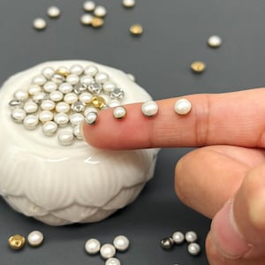 Könnte beinhalten: Eine Sammlung kleiner, runder, perlenartiger Perlen in verschiedenen Metallfassungen, darunter Gold und Silber. Einige Perlen sind auf einer weißen, dekorativen Schale verstreut, andere liegen auf einem Finger. Die Perlen haben eine glänzende, cremefarbene Farbe.