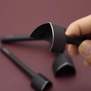 May include: Close-up of several black leather working tools. One tool is held in a hand, showcasing a curved blade with a silver edge. The tools are set against a burgundy background, highlighting their sleek design.