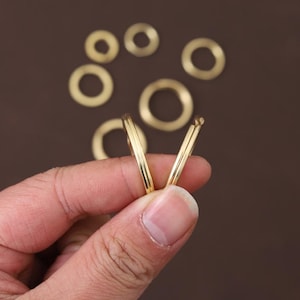 May include: A hand holding two gold hoop earrings, with several other gold rings scattered in the background. The earrings are thin and circular, with a polished finish. The background is a solid brown color.