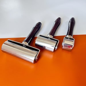 May include: Three leathercraft tools with dark brown wooden handles and shiny metal rollers. The tools are arranged on an orange and white background. The rollers are silver in colour.