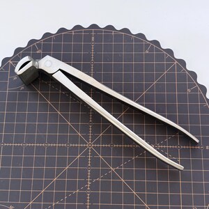 Puede incluir: Una tenaza de herrador de metal plateado, dise&ntilde;ada para recortar cascos de caballos. La herramienta tiene un cabezal de corte y mangos largos para hacer palanca. Se encuentra sobre una esterilla de corte negra.