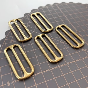 May include: Five gold-coloured rectangular slides with a double-bar design. These metal hardware pieces are arranged on a dark grey cutting mat with white grid lines, suitable for leathercraft or bag making.