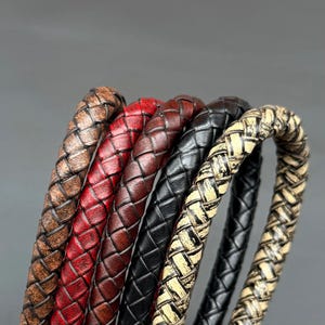 May include: A collection of five braided bracelets featuring distinct textures and colors, including distressed brown, vibrant red, deep burgundy, solid black, and a weathered beige and black pattern, all arranged in a vertical row.