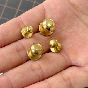 Op de afbeelding: Vier kleine, goudkleurige metalen knopen met een afgeronde bovenkant en een platte basis. De knopen zijn gerangschikt op een lichtgekleurd oppervlak, wat hun gepolijste afwerking en eenvoudige ontwerp laat zien. Deze knopen zijn geschikt voor diverse naai- en knutselprojecten.