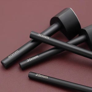May include: A collection of black leathercraft tools, including a mallet and several cylindrical punches. The punches are labeled with sizes: 25mm, 30mm, 35mm, and 40mm. The tools are arranged on a dark red surface.