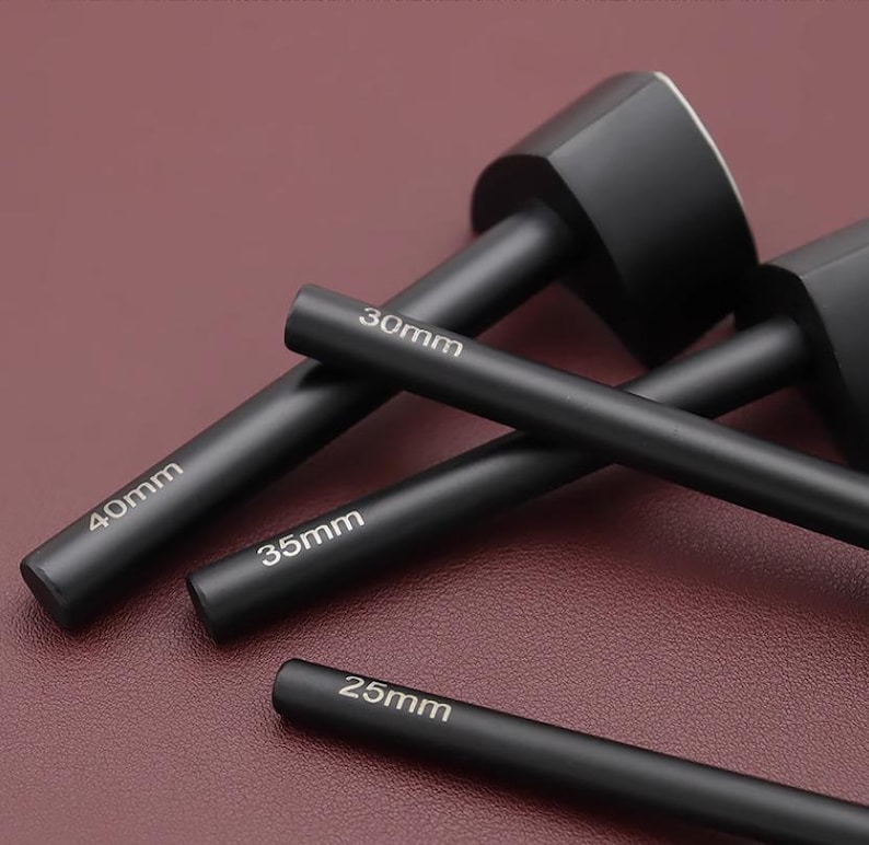 May include: A collection of black leathercraft tools, including a mallet and several cylindrical punches. The punches are labeled with sizes: 25mm, 30mm, 35mm, and 40mm. The tools are arranged on a dark red surface.
