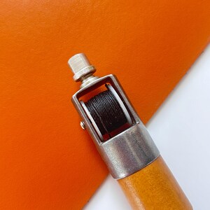 May include: A leathercraft tool with a wooden handle and a metal head holding a spool of black thread. The tool is positioned over a piece of orange leather, showcasing its use in sewing or crafting.