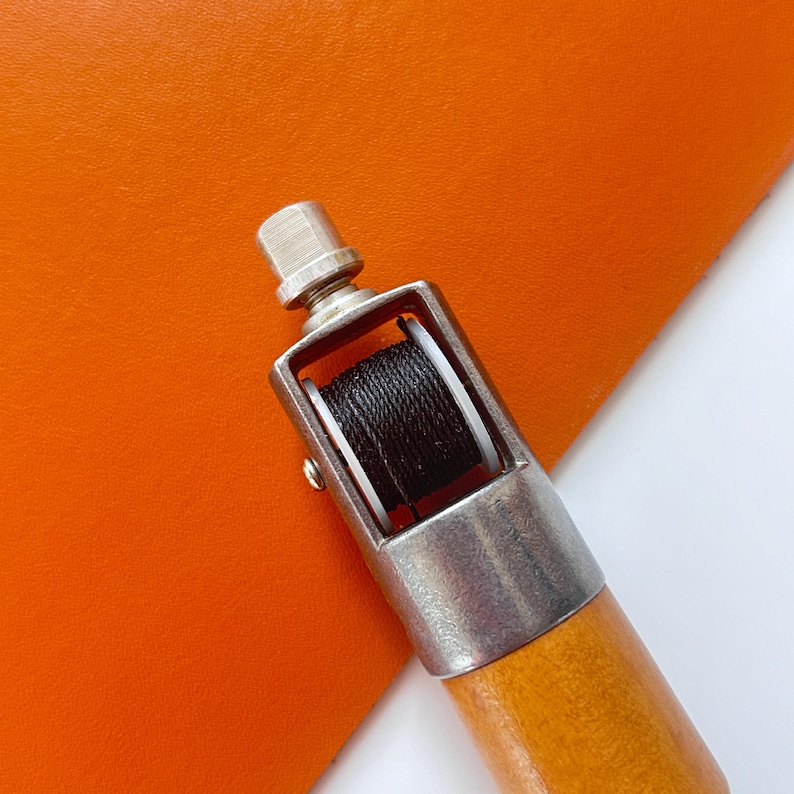May include: A leathercraft tool with a wooden handle and a metal head holding a spool of black thread. The tool is positioned over a piece of orange leather, showcasing its use in sewing or crafting.