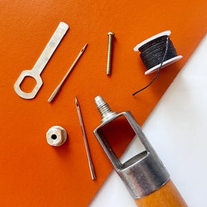 May include: A leathercrafting kit with various tools arranged on an orange surface. The kit includes a metal punch, needles, a spool of black thread, a screw, and a metal tool with a wooden handle. The tools are for leatherwork.