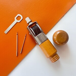 May include: A leathercrafting set with a wooden handle stitching awl, needles, a wrench, and a wooden knob. The awl has a silver metal frame and holds black thread. The tools are arranged on an orange leather surface and a white background.