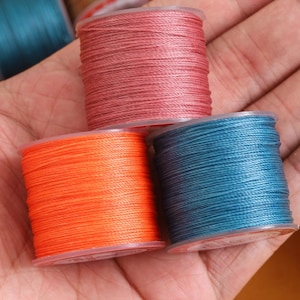 May include: Close-up of several spools of colorful thread. The spools are in shades of orange, teal, and rose. The thread is tightly wound on clear plastic spools. Other spools of thread in shades of brown and teal are visible in the background.