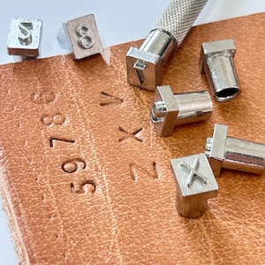 May include: Close-up of metal letter and number stamps with a textured handle, alongside stamped numbers and letters on a piece of brown leather. The stamps include the numbers 5, 8, and the letters V, X, and Z.