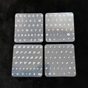 May include: Four clear, square stencils with white lettering and symbols. The stencils include uppercase and lowercase alphabets, numbers, and various symbols. One stencil has the word "Tel:" printed on it.