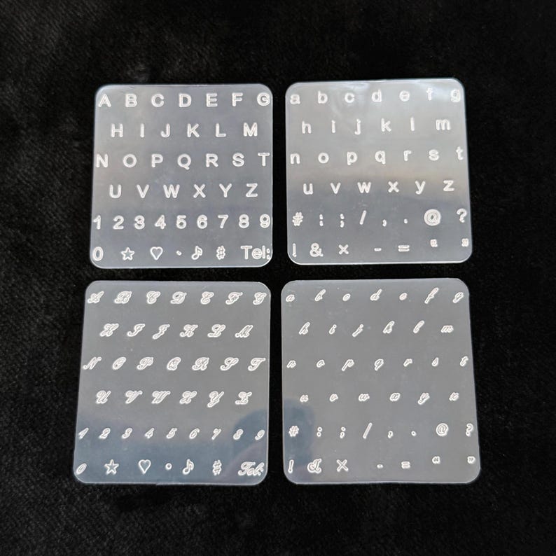 May include: Four clear, square stencils with white lettering and symbols. The stencils include uppercase and lowercase alphabets, numbers, and various symbols. One stencil has the word "Tel:" printed on it.