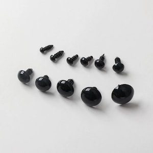May include: A collection of black plastic safety eyes and screw-on fasteners, arranged on a white surface. The eyes are round and glossy, with a screw post for attachment. The fasteners are small, black screws.