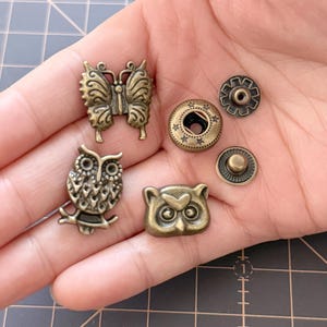 Peut inclure: Un assortiment de boutons-pression et d'ornements en métal couleur laiton antique. L'ensemble comprend un papillon, deux motifs de hiboux et divers composants de boutons-pression. Les pièces sont détaillées avec des motifs complexes et des accents en forme d'étoile.