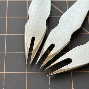 May include: Close-up of three stainless steel leathercraft tools. The tools have a forked design with sharp, pointed tips. The tools are on a dark surface with a grid pattern.