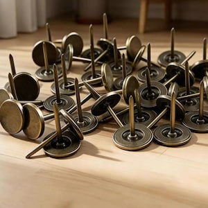 May include: A collection of antique brass upholstery tacks scattered on a wooden surface. Each tack features a round, flat head and a long, pointed pin. The tacks have a vintage, aged appearance, suggesting they are for furniture restoration or decorative projects.