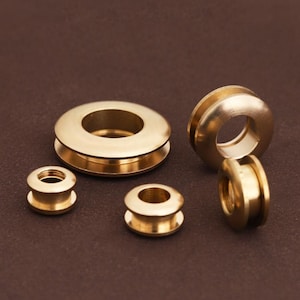 May include: Five brass-colored, circular objects with a hole in the center are arranged on a brown surface. The objects vary in size, with a larger one at the top and smaller ones below. The objects have a shiny, metallic appearance.