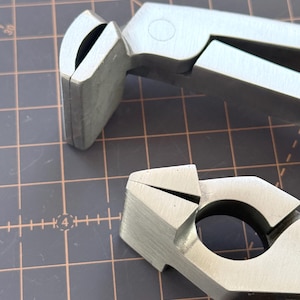 May include: Close-up of two silver metal tools with a matte finish. The tools have a unique design with curved and angled components, suggesting a specialized function. The background is a grid pattern.