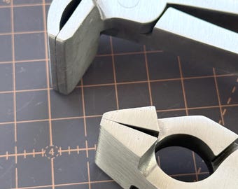 May include: Close-up of two silver metal tools with a matte finish. The tools have a unique design with curved and angled components, suggesting a specialized function. The background is a grid pattern.